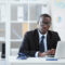 Portrait of confident African manager in eyeglasses and in formal wear looking at camera while sitting at workplace in front of laptop at office