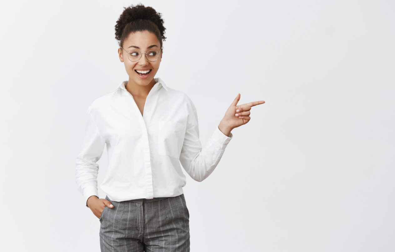 Check it out girl. Portrait of amazed and pleased good-looking happy dark-skinned woman in transparent glasses, holding hand in pocket looking and pointing right with slight smile, being impressed.